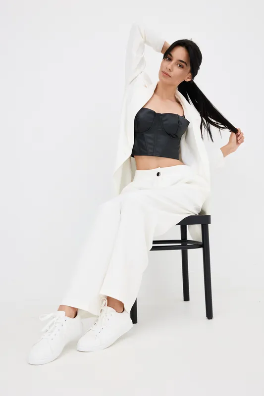 Young woman wearing stylish black top and white suit sitting on chair with playful pose against bright white background, showcasing modern fashion and casual elegance.