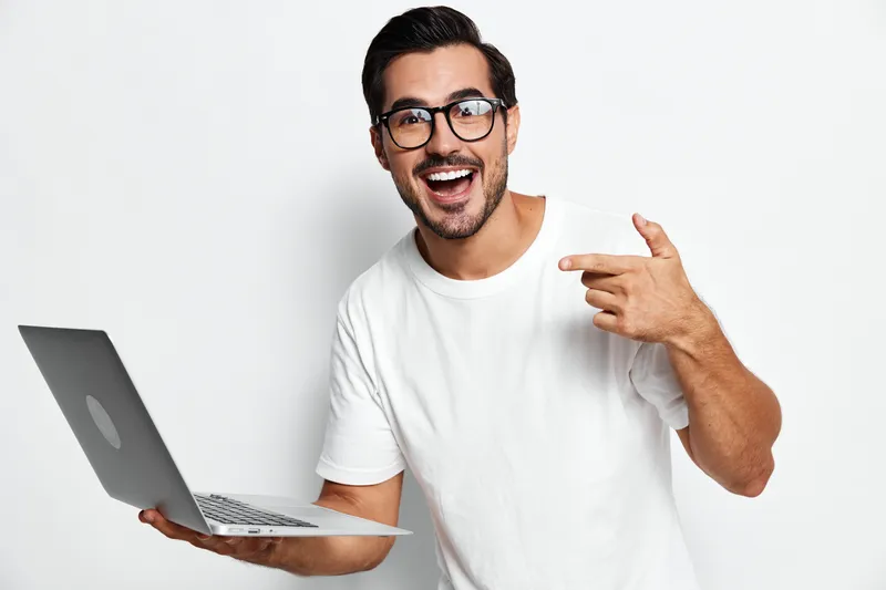 Smiling man wearing white t-shirt holding laptop while pointing finger at camera with cheerful expression, isolated on plain solid background. Tech lifestyle concept.