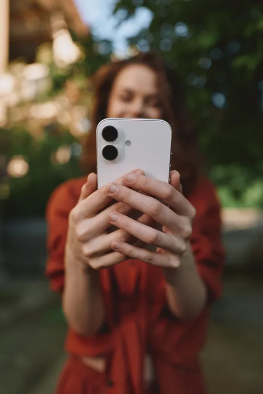 Smiling woman in casual outfit taking a closeup selfie outdoors with a smartphone. Natural light, green background, lifestyle moment focused on self expression and everyday connection