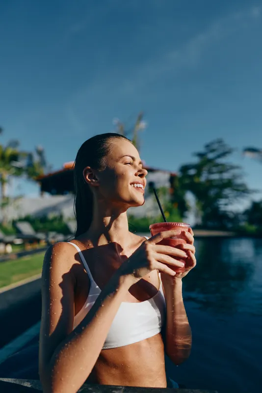 Cheerful woman outdoors by a pool, enjoying a refreshing drink, sunlit moment, relaxed posture, warm breeze, vibrant colors, tropical backdrop, lifestyle mood