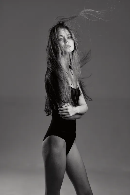 Black and white portrait of a confident woman with long hair in a bodysuit posing against a simple background