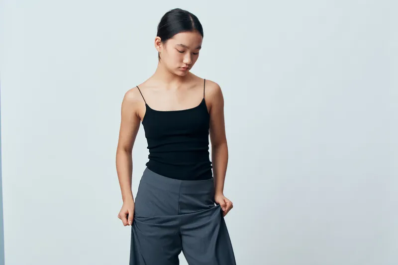 Young woman wearing black tank top and gray pants standing against plain background looking down with thoughtful expression and loose hair tied back in casual style.