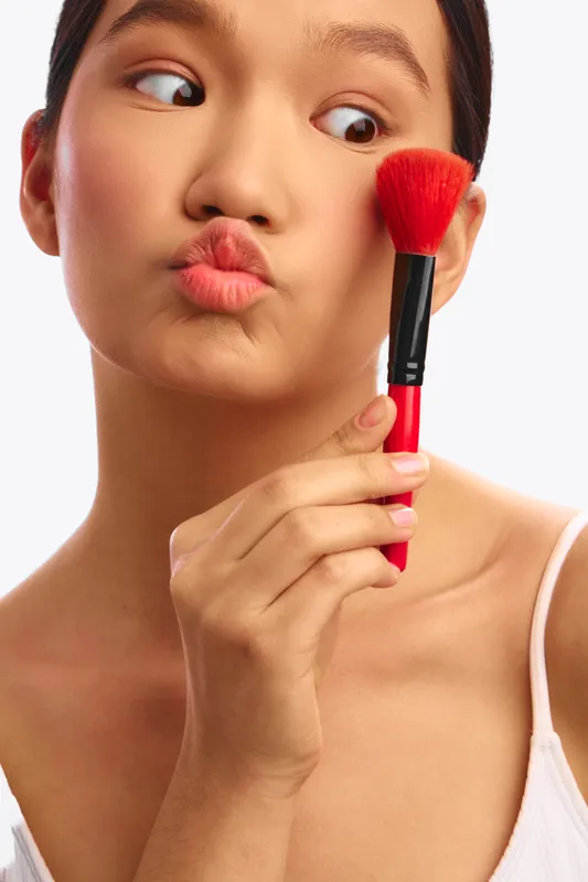asian woman smiles softly while applying makeup with a brush, captured in a close up that emphasizes focus, style, and beauty care in a bright studio setting.