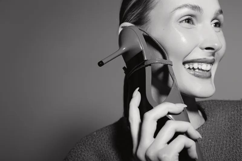 Smiling Caucasian woman with light skin holds high heel shoe near ear as if talking on phone, close-up in black and white. Concept for creativity, communication, and playful moments.
