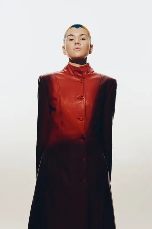 Woman wearing a dark red high-collared coat standing confidently against a light neutral background, showcasing modern fashion and minimalistic style.