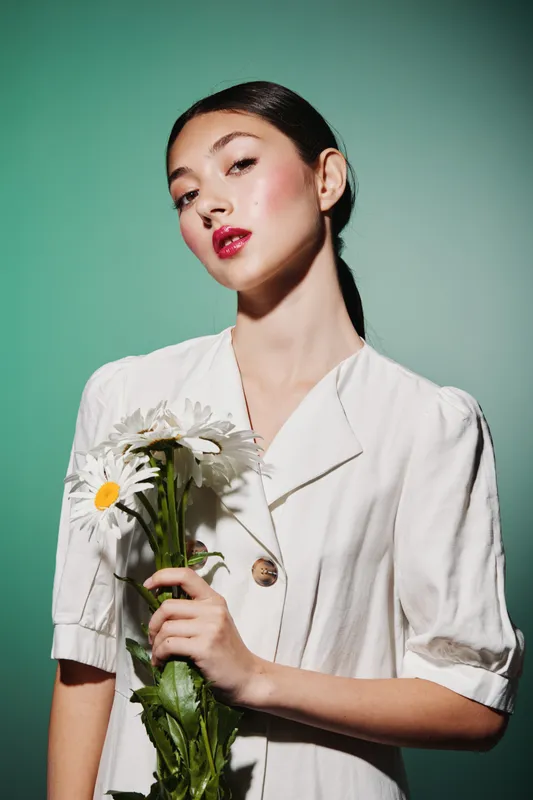 Young woman wearing a white outfit holding white flowers, showcasing glossy skin and bold glossy lips on a bright green background. Concept for fashion beauty and skincare.