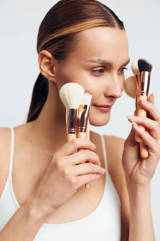 Young woman with clean skin holding makeup brushes near her face on a white background. Concept of beauty, skincare, and natural makeup application with soft lighting.