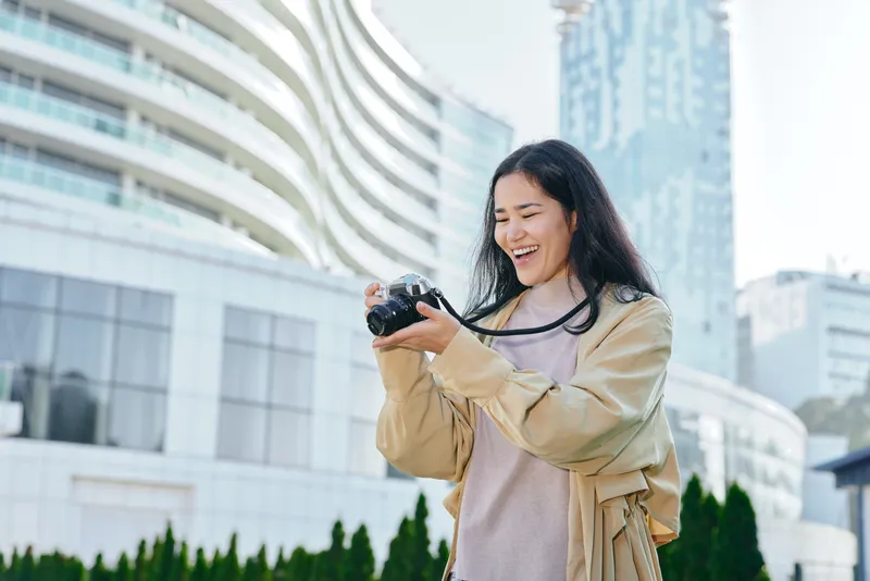 Smiling woman holds a camera outdoors in an urban setting, capturing moments among buildings and greenery, casual style, lifestyle photography for travel, urban life and everyday moments.