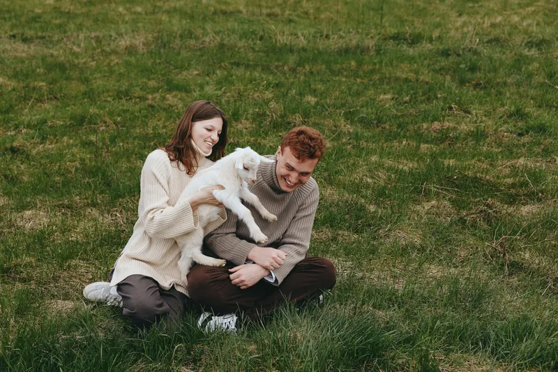 Young couple sitting on grass in casual clothes enjoy playful moment with a white baby goat in a natural outdoor setting during springtime.