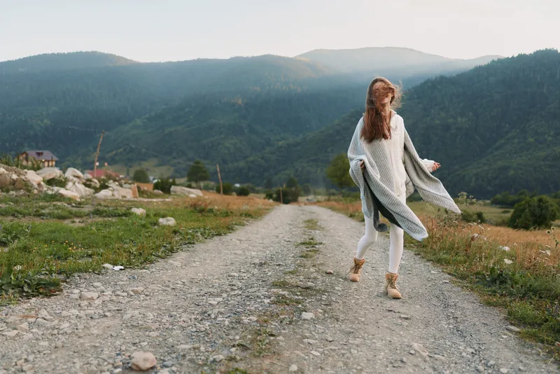 Young woman in cozy sweater walking barefoot on rural gravel road with mountains in background enjoying peaceful lifestyle and fresh air outdoors.