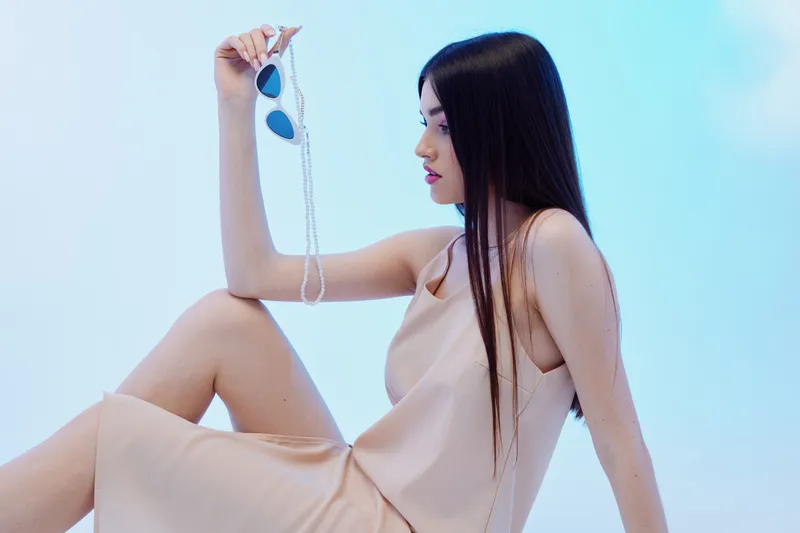 Young woman in a beige dress holds sunglasses with a pearl chain, sitting against a light blue background. Concept of beauty, fashion, and elegance with a calm mood.