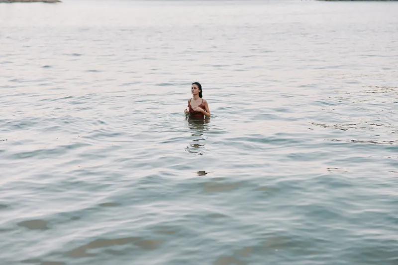 Solitary figure stands waist deep in calm water, sunlight softly highlighting the shoulders and gentle ripples. A quiet moment of reflection in open, tranquil water.