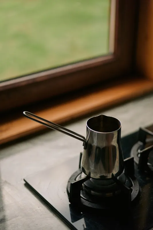 Coffee pot on a stove beside a window in a warm kitchen scene, a metal cezve ready for brewing, captured in soft natural light.