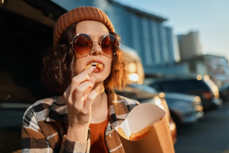 Woman eating fries in sunglasses and beanie near car on city street at golden hour, candid moment with authenticity, candid lifestyle, golden hour glow, mindful living and emotional storytelling.