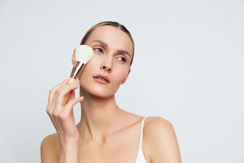 Young woman applying powder with brush on clean face, minimal makeup, natural skin tone, soft lighting, plain background, beauty routine, skincare focus