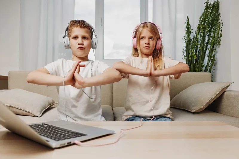 Two children wearing headphones practice yoga poses at home sitting on sofa in bright living room with laptop on table. Concept for mindfulness, wellness, and healthy lifestyle.