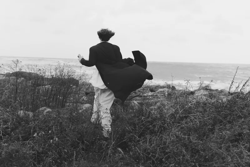 Couple in coats move through a windy coastal field, cloak billowing as they walk toward the sea, capturing motion, freedom and quiet embrace in a stark black and white landscape