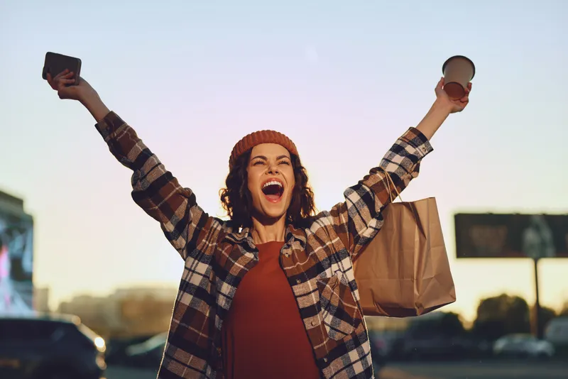 Woman joy coffee shopping golden urban candid authenticity lifestyle glow mindful living emotional storytelling portrait of a smiling woman with arms raised holding takeaway and paper bag at sunset