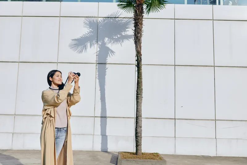 Woman with camera photographing a palm tree and white wall in an outdoor urban setting, casual outfit, candid moment of exploration and creative photography