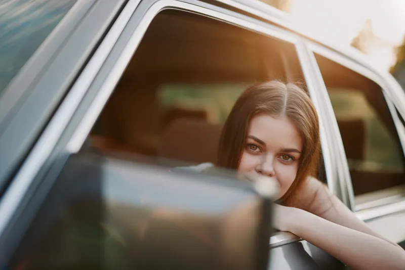 Young woman with long hair enjoying a sunset view from a car window, evoking feelings of freedom and adventure, dressed casually in a summer outfit.