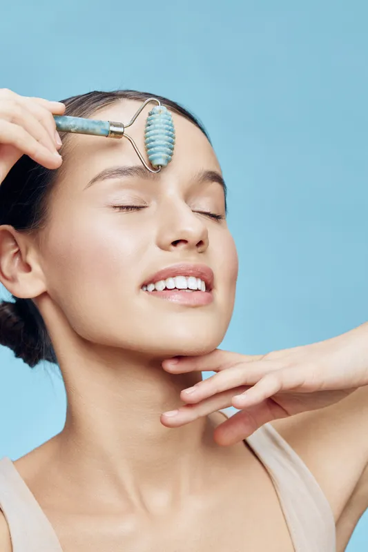 Young woman performing facial massage with a jade roller, enhancing skincare routine, glowing skin, beauty concept, light blue background, wellness vibe