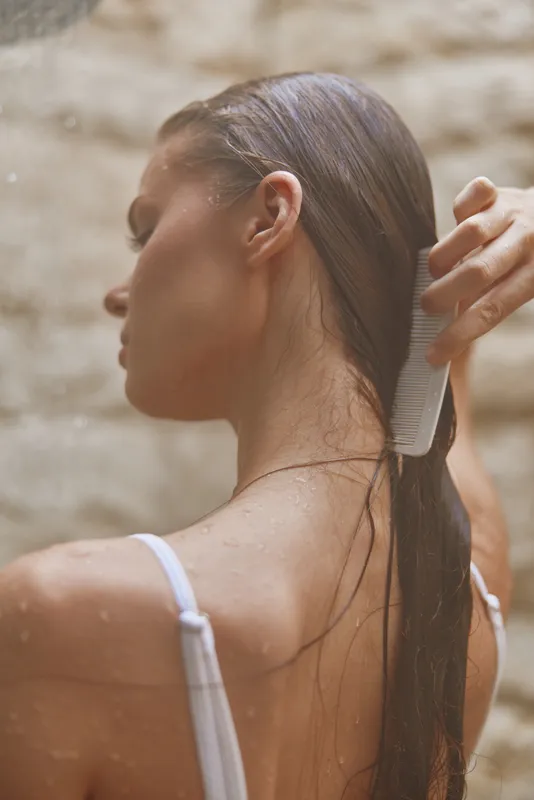 young woman combing wet hair with white comb, natural skin, spa wellness, soft sunlight, calm mood, close-up back view