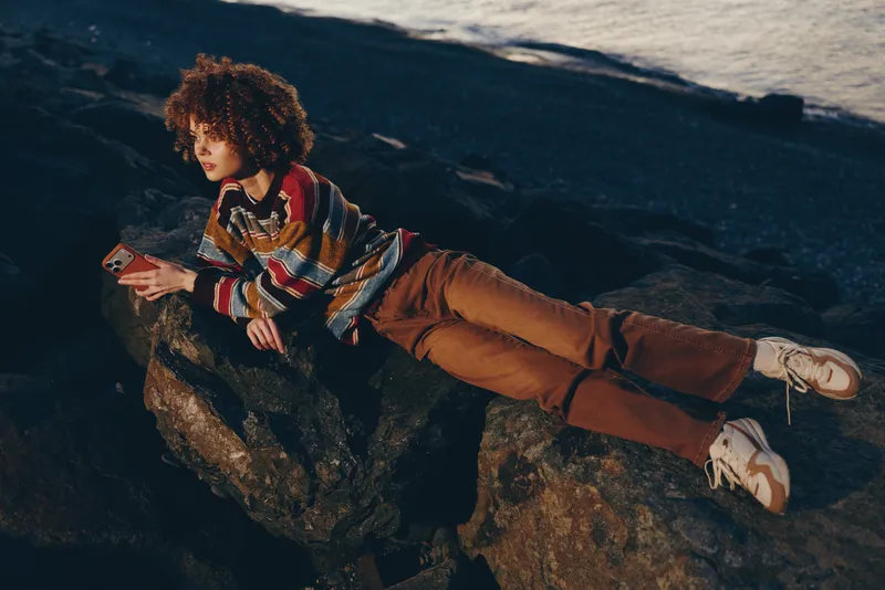 Woman lifestyle outdoors rock scene, curly hair fashion, holding smartphone while relaxing on rugged stone at dusk, casual travel mood
