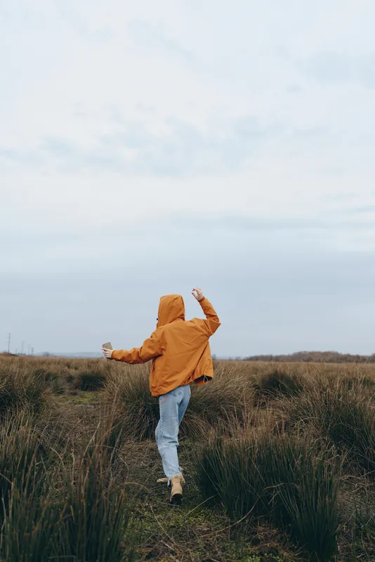 Woman outdoors in a grassy field, walking forward with cheerful posture, wearing orange jacket and jeans, windy breeze, cloudy sky, natural light, lifestyle moment, soft motion, fresh air