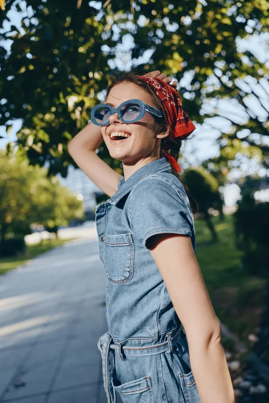 Lifestyle woman in modern boho-western denim street style with sunglasses and a red bandana smiling joyfully outdoors, captured with warm film color tones.