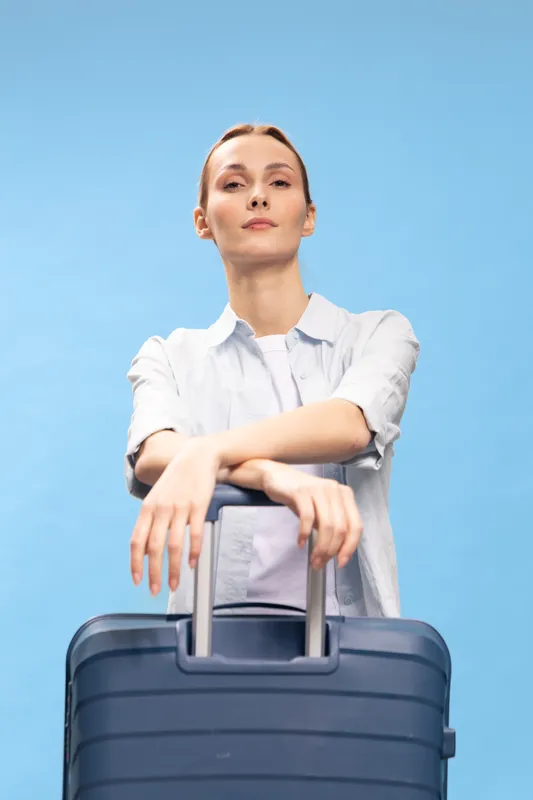 Confident young woman in casual clothes stands with suitcase handle in front of light blue background. Concept for travel, vacation, and modern lifestyle with calm expression and relaxed pose.