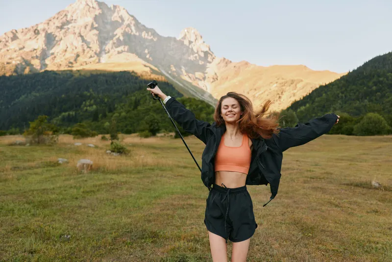 A cheerful woman stands outdoors in a rugged mountain landscape, arms raised in joy, wearing a coral crop top, black jacket, and shorts, smiling brightly in warm sunlight.