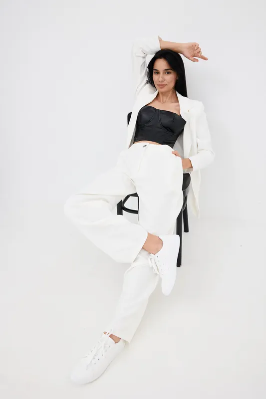 stylish woman sitting casually in white pants and blazer with black corset top, relaxed pose against white background, modern fashion and minimalism