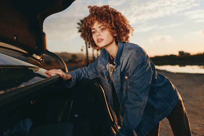 Woman lifestyle by car, curly hair woman in denim jacket leaning on open door at sunset outdoors, relaxed smile, warm light, countryside road background, casual fashion