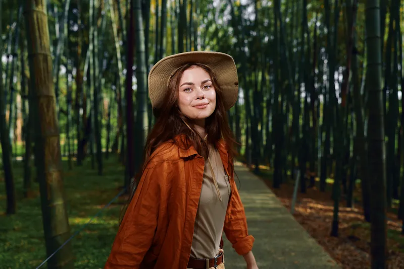 Woman in bamboo forest wearing straw hat and orange jacket walking on wooden boardwalk path, smiling at camera. Nature portrait, travel, leisure, peaceful getaway and outdoor lifestyle.