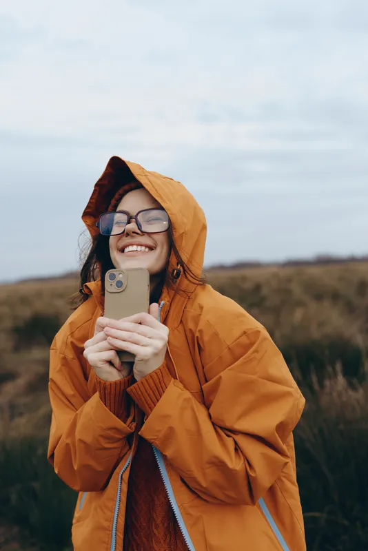 Woman lifestyle outdoors, orange raincoat with hood, smiling while holding phone, countryside background with wind and clouds, concept of communication and connection for everyday confidence