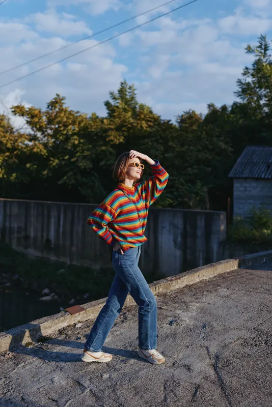 Young woman in a colorful striped sweater and blue jeans walks outdoors in a rural setting, shielding her eyes with sunglasses. Casual outfit, bob haircut, relaxed travel and lifestyle portrait in