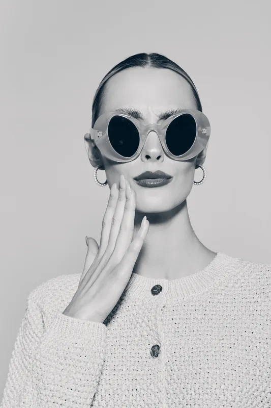 Fashionable white woman with pale skin wearing large round sunglasses and a knitted sweater touches her face in stylish black and white portrait.