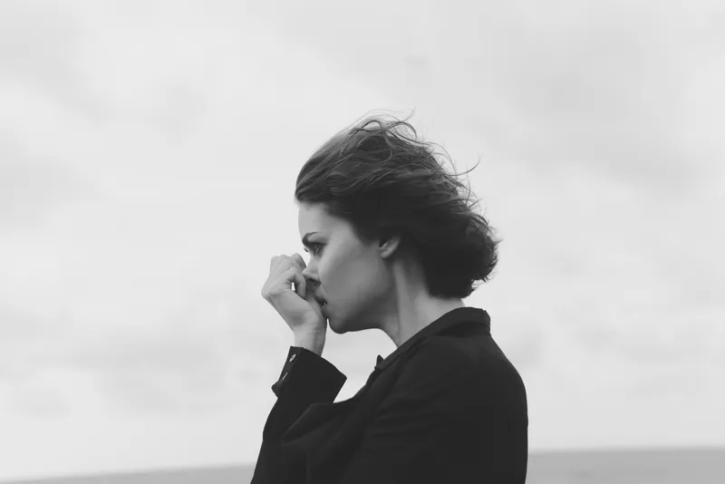 Monochrome image of a contemplative woman in a dark coat by the sea, windblown hair and a distant horizon, conveying solitude, mood, minimalist fashion and a dramatic yet calm outdoor scene