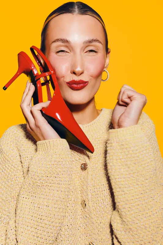 Smiling woman in beige sweater holds red high heel shoe near face with closed eyes on yellow background. Concept of fashion, style, footwear, and joyful emotions.