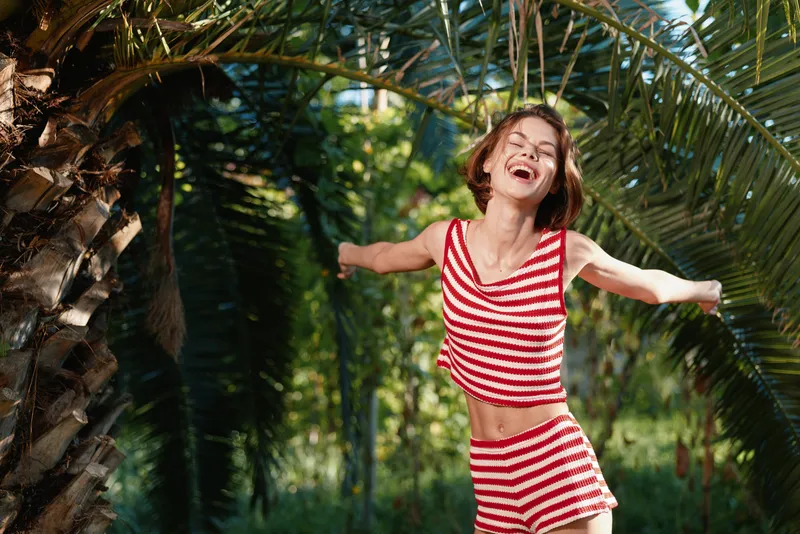 A cheerful woman wearing a red and white striped outfit enjoys a sunny outdoor moment among lush tropical plants, expressing freedom, joy, and playful energy.