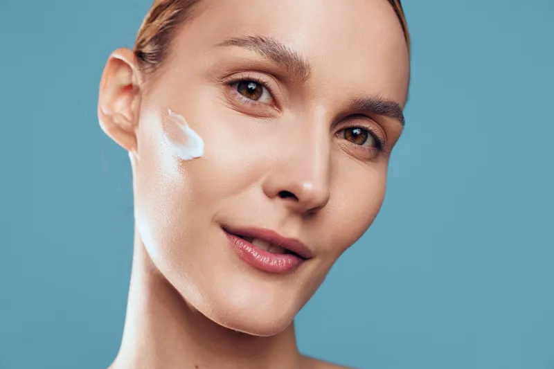 A young woman applies cream to her cheek in a bright studio, illustrating a focused skincare routine and a quiet moment of self care amidst soft, even lighting.
