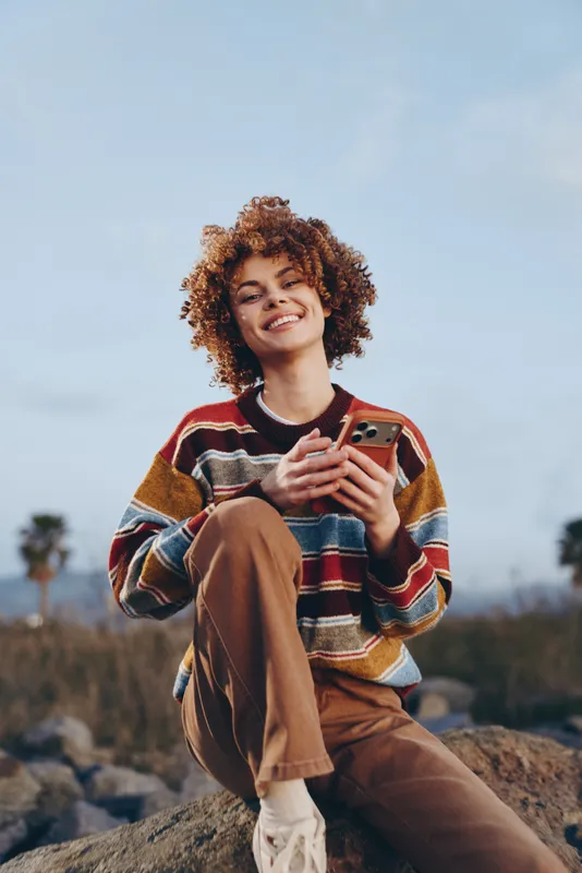 Woman lifestyle outdoors in field under blue sky, curly hair young adult seated on rock, wearing striped sweater, holding phone, relaxed smile, casual leisure portrait in natural landscape daylight