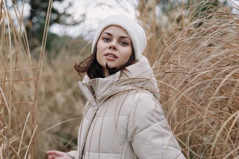 Portrait of a woman in a light beige coat and white beanie outdoors among tall dry grasses, capturing winter fashion and a serene natural mood.