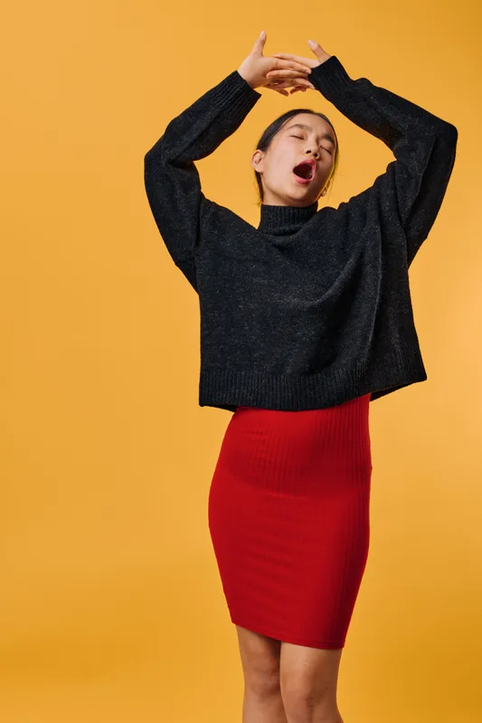 Woman in black sweater and red skirt yawning with raised arms against yellow background. Casual fashion, tired gesture, female model stretching and waking up in studio portrait.