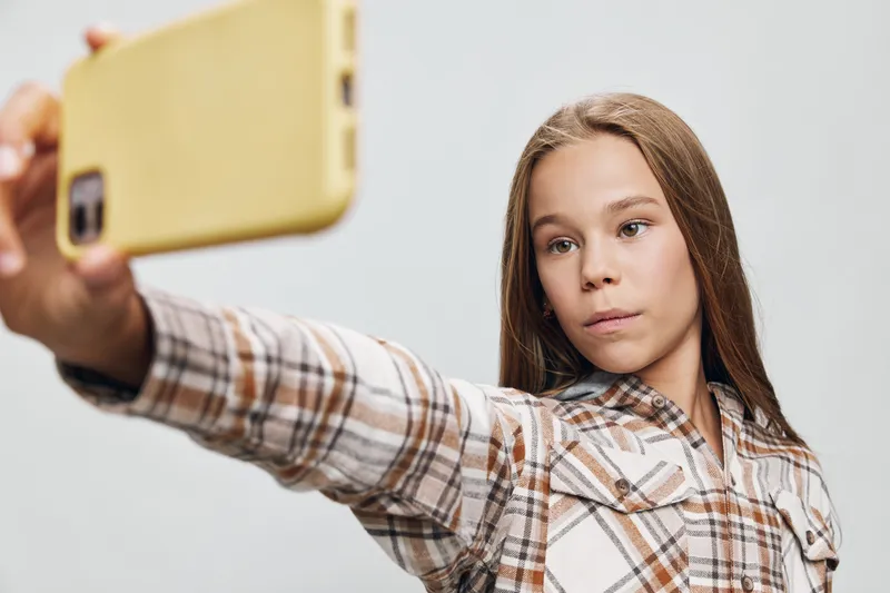 Teenage girl with long brown hair taking selfie on yellow smartphone isolated on white background wearing plaid shirt with focused expression.