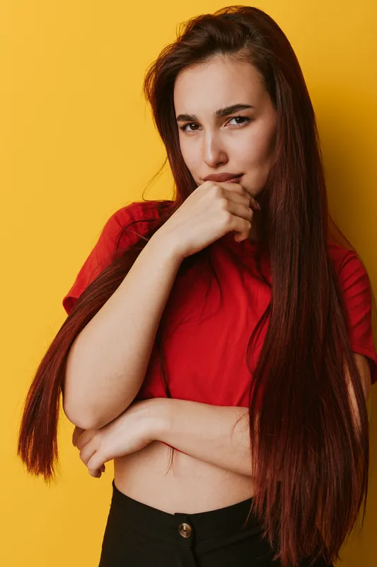 Young woman with long red hair and red shirt posing with thoughtful expression and hand near mouth against yellow background. Casual style and confident mood in studio portrait.