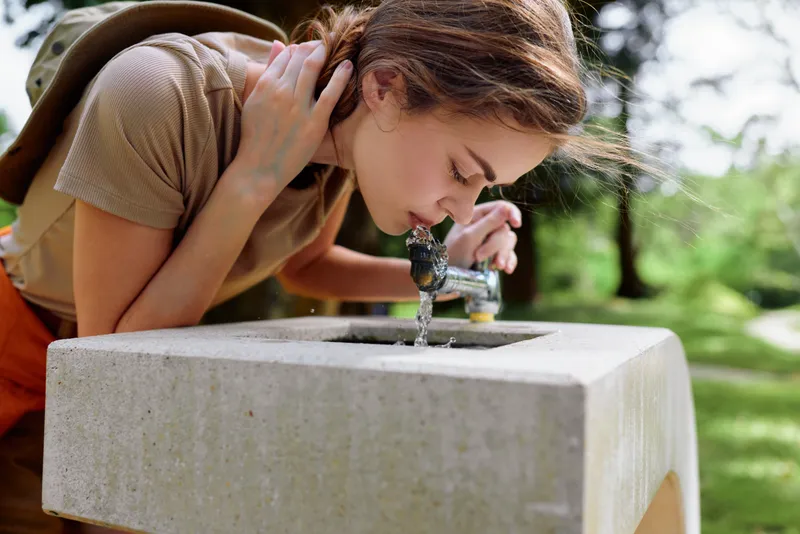 Woman drinking from a public fountain in a park, leaning over with backpack and casual clothes, summer hydration shot capturing profile closeup and natural green outdoor setting.