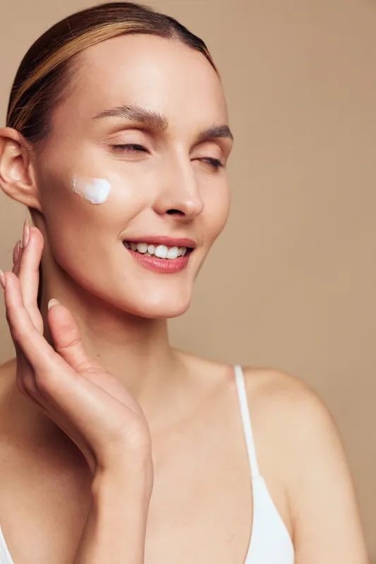 A relaxed woman applies cream to her cheek with a gentle smile, revealing a simple skincare moment and a healthy, hydrated complexion in a soft, warm studio.