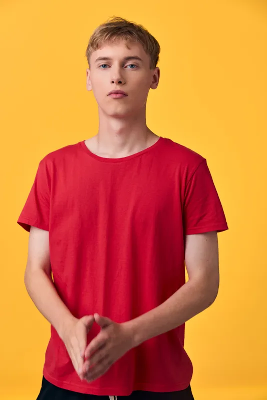 Red shirted young man stands in a studio against a bright yellow backdrop, hands clasped in front, neutral expression, bold minimal shot ideal for fashion, lifestyle or advertising use