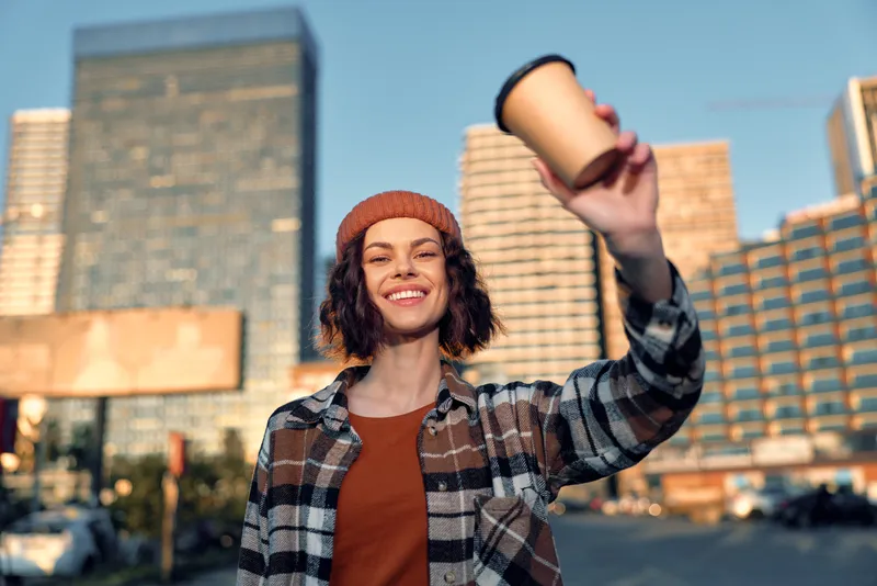 woman holding coffee cup in city street, smile and urban vibe under golden light; candid lifestyle portrait with authenticity, golden hour glow, mindful living and emotional storytelling